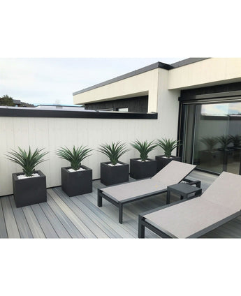 Een moderne patio heeft twee grijze loungestoelen en vijf vierkante plantenbakken, elk met een Kunstplant Agava Pandora UV 85 cm, tegen een wit met zwart gepaneelde muur voor een strakke, minimalistische look.