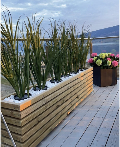 Ein Pflanzgefäß aus Holz mit grünem UV-Kunstrasen (120 cm) und weißen Steinen steht auf einem Balkon mit Glasgeländer. In der Nähe überblickt ein schwarzer, quadratischer Topf mit blühenden rosa, grünen und weißen Hortensien die malerische Aussicht.