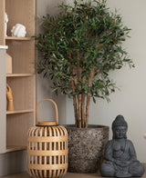 Een Kunst olijfboom UV 120 cm staat naast een grijs Boeddhabeeld en een houten lantaarn op de vloer, naast een lichte houten plank met handgevormde ornamenten. Deze onderhoudsarm plant brengt een rustige, serene sfeer in de ruimte.
