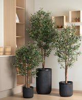 De kunst olijfboom UV 120 cm staat op een lichte houten vloer, met moderne licht houten planken en kasten op de achtergrond. De onderhoudsarme, UV-bestendige zwarte ribbelpot zorgt voor een eigentijdse look.