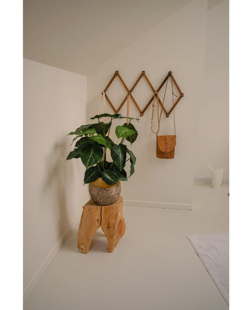 Eine minimalistische Raumecke wird zu einer grünen Oase mit der Kunstpflanze Syngonium UV 58 cm auf einem Hocker aus Naturholz, neben einer braunen Ledertasche auf einem geometrischen Wandregal und einem weißen Krug, alles vor cremefarbenen Wänden und Boden.