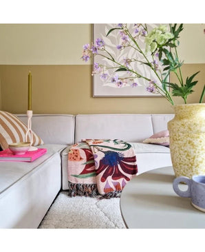 Een gezellige woonkamer met een lichtbeige sofa, een kleurrijke deken met bloemen en kussens met een patroon. Op een ronde tafel staan een gele vaas met Kunsttak Daphne Spray 108 cm paars en een klein blauw kopje. Aan de neutraal getinte muur hangt kunst.