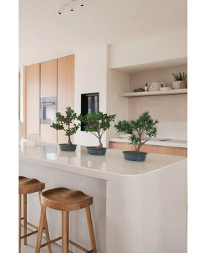 Een Kunstplant Bonsai Juniper 40 cm staat op een wit keukeneiland met twee houten krukjes, omringd door lichthouten kastjes, witte werkbladen en een minimalistische inrichting.
