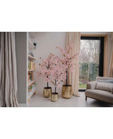 Een Kunstplant Bougainvillea 120 cm roze staat bij het raam in een gezellige woonkamer met witte muren, een boekenplank, beige gordijnen en een grijze fauteuil.