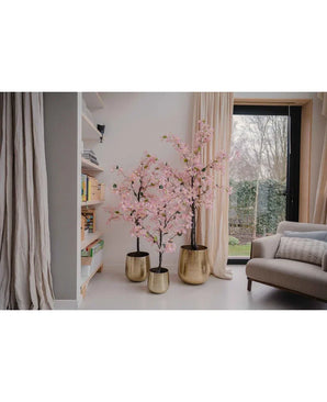 Een Kunstplant Bougainvillea 180 cm roze staat in een gouden plantenbak bij een raam in een moderne woonkamer, naast een boekenplank en beige gordijn, met rechts een grijze fauteuil zichtbaar.