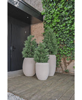 Drie grote beige plantenbakken met Kunstplant Conifeer deluxe UV 150 cm staan op een houten vlonder bij een zwarte deur en klimop muur, waardoor een stijlvolle buiteningang of patio ontstaat voor iedereen die kunstplant tuin inspiratie zoekt.