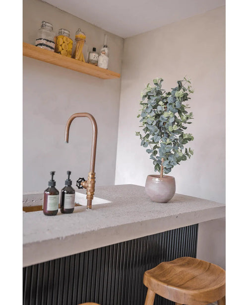 Eine moderne Arbeitsplatte verfügt über einen goldenen Wasserhahn, drei Seifenspender, die künstliche Eukalyptuspflanze (91 cm) und Holzhocker. Minimalistische Regale über den Gläsern mit verschiedenen Zutaten sorgen für eine gemütliche Atmosphäre.
