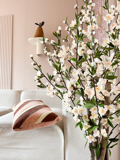 Ein gemütliches Wohnzimmer mit einem weißen Sofa, einem gestreiften rosa-braunen Kissen und dem 120 cm großen cremefarbenen Blütenzweig der künstlichen Kirschblüte im Vordergrund. Eine Topfpflanze steht auf einem Wandregal vor einer blassrosa Wand.