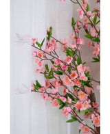 De Kunstplant Kersenbloesem 120 cm roze, met roze kersenbloesem en groene bladeren, staat naast een doorschijnend wit gordijn, de bloemen zacht zichtbaar door de doorschijnende stof.