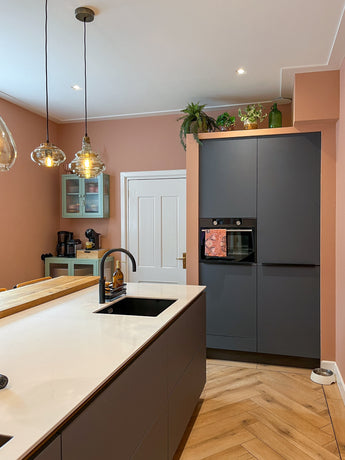 Moderne keuken met perzikkleurige muren, donkergrijze kasten, wit aanrechtblad, hanglampen en visgraatmotief. Een Kunstplant Monstera 18 cm en glazen flessen bovenop de kasten zorgen voor een frisse touch.