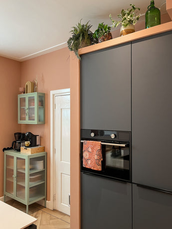 Een moderne keuken met matte donkere kasten, een ingebouwde oven en een handdoek met patroon. Bovenop de kasten staan potplanten zoals Kunstplant Monstera 18 cm en glazen flessen, terwijl een mintgroene kast in de hoek koffiebenodigdheden bevat.
