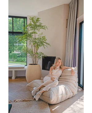Een vrouw in lichte casual kleding ontspant op een crèmekleurige loungestoel in een moderne woonkamer met houten vloeren, grote ramen, natuurlijk licht en een Kunstplant Ruscus 180 cm die groen toevoegt aan de ruimte.