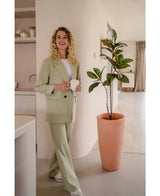 Een glimlachende vrouw met krullend blond haar, gekleed in een lichtgroen pak en witte sneakers, staat binnen met een witte mok naast de Kunstplant Yulaniaboom 120 cm in een moderne, neutraal gekleurde kamer.