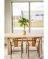Een lichte houten tafel met twee bijpassende stoelen staat op een rond kleed bij een groot raam. Een Kunstplant citroenboom 75 cm siert het midden van de tafel en voegt charme toe aan het lichte, minimalistische tafereel.
