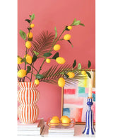 Vor einer korallenrosa Wand stehen eine gestreifte Vase mit Kunsttak-Zitronenzweigen von 104 cm in Gelb, ein farbenfrohes Gemälde, ein Stapel Bücher, ein blauer Kerzenständer mit einer Kerze und ein Tablett mit Zitronen.