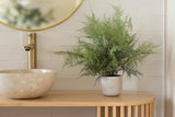 De Kunstplant Asparagus 43 cm in sierpot staat op een houten aanrecht naast een ronde stenen wastafel en gouden kraan, met een ronde spiegel die het tafereel weerspiegelt tegen een wit betegelde muur.