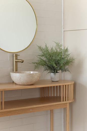 Een minimalistisch badkamermeubel heeft een ronde spiegel, een gouden kraan, een stenen wasbak en de Kunstplant Asparagus 43 cm in sierpot op een licht houten aanrechtblad tegen wit betegelde muren.