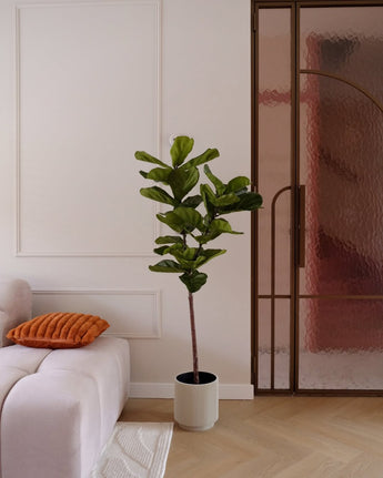 Een Kunstplant Ficus Tabaksplant Deluxe 155 cm in een witte pot staat naast een lichtroze bank met een oranje kussen, bij een glazen deur met een bronzen frame, in een moderne minimalistische woonkamer.