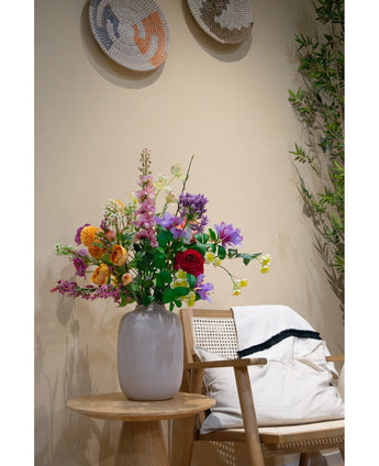 Een witte vaas met vijf levendige oranje Ranonkel kunsttak x5 Real Touch 62cm bloemen staat op een houten tafel naast een houten stoel met een wit kussen en een deken; gevlochten manden hangen aan een beige muur en groene takken gluren dichtbij.